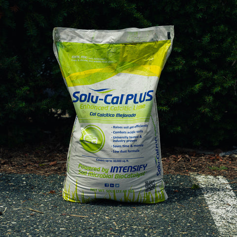 A large bag of Solu-Cal Plus Enhanced Calcitic Lime fertilizer, featuring a white and green design with blue text, standing on a paved surface with dark green foliage in the background.
