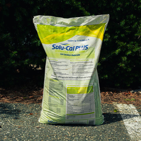 A large white and yellow bag of Solu-Cal Plus Enhanced Calcium Lime fertilizer stands on asphalt outdoors with dark green foliage in the background.