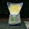 A 50 lb bag of Solu-Cal USA Humic PLUS soil conditioner stands on asphalt in front of a dark green pine hedge. The bag is white with blue graphics and a yellow informational label on the upper portion.