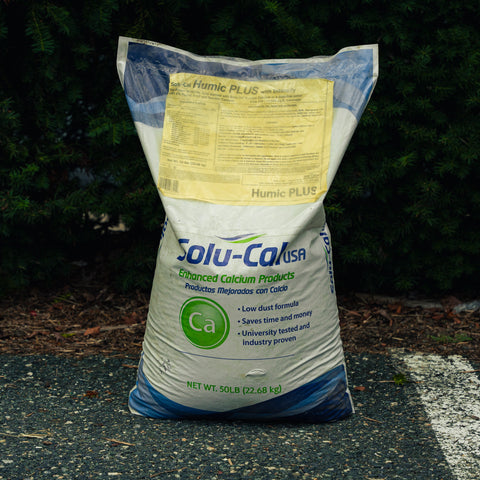 A 50 lb bag of Solu-Cal USA Humic PLUS soil conditioner stands on asphalt in front of a dark green pine hedge. The bag is white with blue graphics and a yellow informational label on the upper portion.