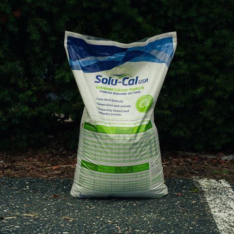 A large white bag of Solu-Cal USA Enhancing Calcium Products standing upright on asphalt in front of a dark green hedge. The bag is blue, white, and green, with the brand name 'Solu-Cal USA' clearly visible.