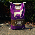 A large purple and brown bag of Country Acres Horse Feed standing upright on asphalt outdoors with bushes in the background.