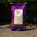 A large purple and brown bag of Country Acres Horse Feed standing upright on asphalt outdoors with bushes in the background.