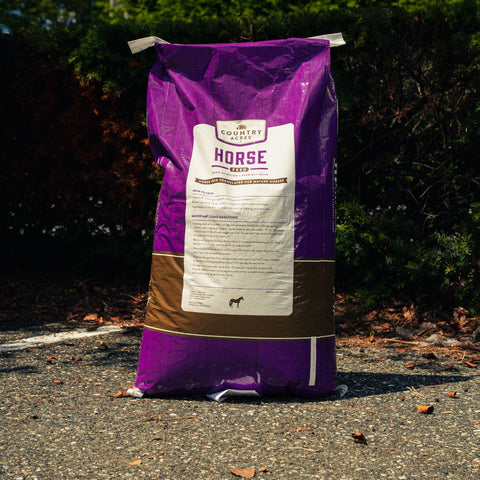 A large purple and brown bag of Country Acres Horse Feed standing upright on asphalt outdoors with bushes in the background.