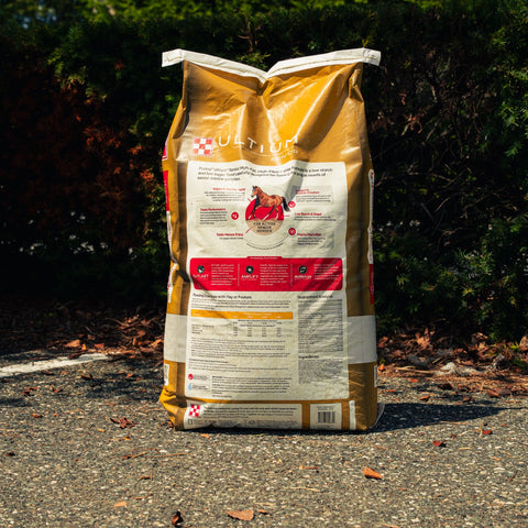 A large bag of Purina Ultium Senior horse feed supplement stands on a gravel path outdoors with dark green bushes in the background. The bag is tan and white with a photo of a brown horse and the Purina logo.