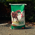 A large bag of Purina 300 Omolene Mare & Foal horse food, standing upright on an asphalt surface outdoors with green foliage in the background.