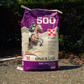A large bag of Omolene 500 Competition horse feed supplement, shown standing upright on an asphalt surface with a blurred outdoor background of green foliage.