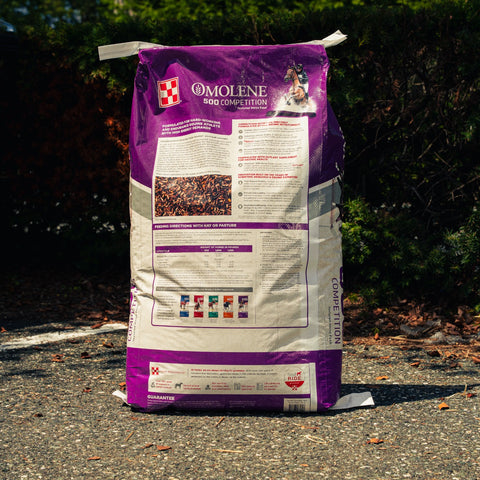 A large bag of Omolene 500 Competition horse feed supplement, shown standing upright on an asphalt surface with a blurred outdoor background of green foliage.