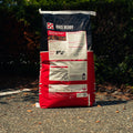 A 50 lb bag of Purina Race Ready horse feed supplement stands on an asphalt ground with dark green foliage in the background. The bag is red, black, and white with a graphic of a horse and jockey.