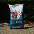 A large bag of Purina Enrich+ Senior horse feed supplement stands on an outdoor gravel path with dark green bushes in the background. The bag is white and blue with a photo of a brown horse on the front and the Purina brand logo.
