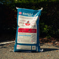A large bag of Purina Enrich+ Senior horse feed supplement stands on an outdoor gravel path with dark green bushes in the background. The bag is white and blue with a photo of a brown horse on the front and the Purina brand logo.