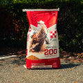 A large red and white bag of 200 Omolene Performance horse feed stands upright on an asphalt surface outdoors with dark green foliage in the background.