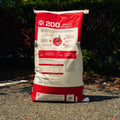 A large red and white bag of 200 Omolene Performance horse feed stands upright on an asphalt surface outdoors with dark green foliage in the background.
