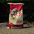A large red and white bag of Purina 200 Omolene Performance RT Horse food stands upright on an outdoor gravel path with dark green foliage in the background.