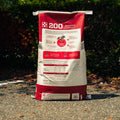 A large red and white bag of Purina 200 Omolene Performance RT Horse food stands upright on an outdoor gravel path with dark green foliage in the background.