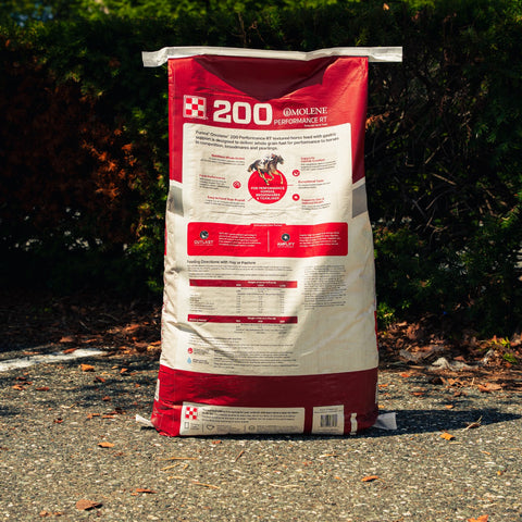 A large red and white bag of Purina 200 Omolene Performance RT Horse food stands upright on an outdoor gravel path with dark green foliage in the background.