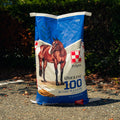 A large bag of Purina Molene 100 Active Pleasure horse feed standing on pavement outdoors with trees and shrubs in the background.