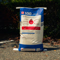 A large bag of Purina Molene 100 Active Pleasure horse feed standing on pavement outdoors with trees and shrubs in the background.