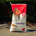 A large bag of Purina Strategy horse feed supplement stands outdoors on a gravel path with dark green foliage in the background. The bag is white and red with a photo of a brown horse on the front and the Purina brand logo.