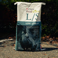 A large bag of Purina L/S Low Starch horse feed standing outdoors on pavement with dark foliage in the background.