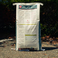 A large white bag of Purina L/S horse supplement stands upright on a gravel surface outdoors with dark green foliage in the background.