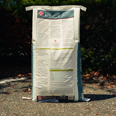 A large white bag of Purina L/S horse supplement stands upright on a gravel surface outdoors with dark green foliage in the background.
