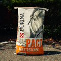 A large bag of Purina Impact Hay Stretcher pelleted horse feed standing upright outdoors on a paved ground with dark foliage in the background.