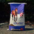 A large bag of Purina Strategy Healthy Edge horse feed supplement stands on asphalt outdoors with dark foliage in the background. The bag is blue and white with a photo of a brown horse and the Purina logo.