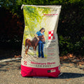 A large bag of Purna Miniature Horse and Pony feed standing outdoors on a gravel path with trees in the background. The bag is off-white with a red bottom section, featuring an illustration of a woman petting a brown pony.