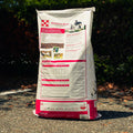 A large bag of Purna Miniature Horse and Pony feed standing outdoors on a gravel path with trees in the background. The bag is off-white with a red bottom section, featuring an illustration of a woman petting a brown pony.