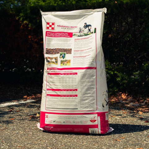 A large bag of Purna Miniature Horse and Pony feed standing outdoors on a gravel path with trees in the background. The bag is off-white with a red bottom section, featuring an illustration of a woman petting a brown pony.