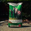 A large green bag of Purina Ultium Growth horse feed stands outdoors on a gravel path with dark green foliage in the background. The bag features photos of a horse and foal and the Purina brand logo.