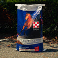 A 50 lb bag of Purina Ultium Competition horse feed stands on asphalt outdoors with trees in the background. The bag is blue and white with a picture of a brown horse and the Purina logo.