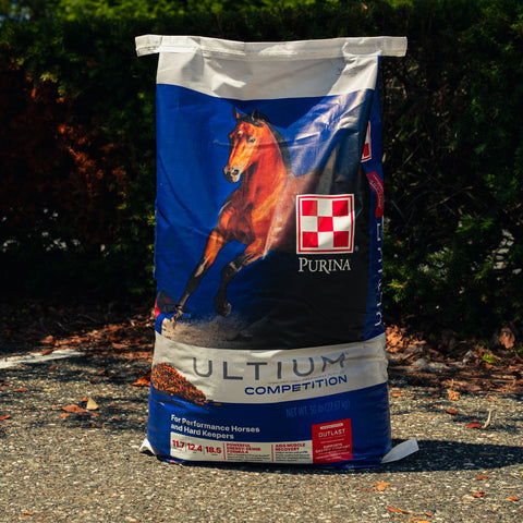 A 50 lb bag of Purina Ultium Competition horse feed stands on asphalt outdoors with trees in the background. The bag is blue and white with a picture of a brown horse and the Purina logo.