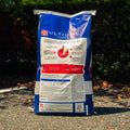 A large blue bag of Ultium Competition horse food stands upright on an outdoor gravel path with dark green foliage in the background.