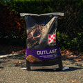 A 40 lb bag of Purina Outlast Gastric Support Supplement for horses, standing on an asphalt surface outdoors with bushes in the background.