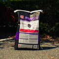 A large bag of Outlast horse food placed on asphalt outdoors with trees and bushes in the background. The bag is mostly black with white, purple, and red label panels.