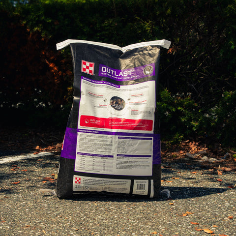 A large bag of Outlast horse food placed on asphalt outdoors with trees and bushes in the background. The bag is mostly black with white, purple, and red label panels.