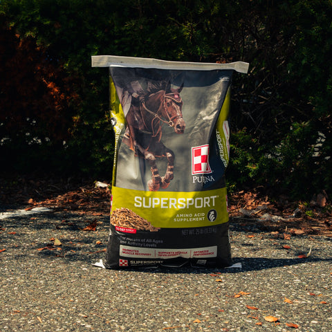 A bag of Purina Supersport horse supplement standing upright on an outdoor paved surface with trees and shrubs in the background.