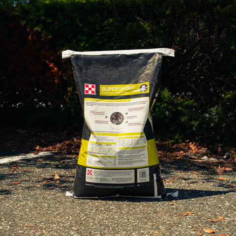 A large black bag of pet supplement powder labeled 'SUPERSPORT' stands upright on an outdoor paved surface with bushes in the background.