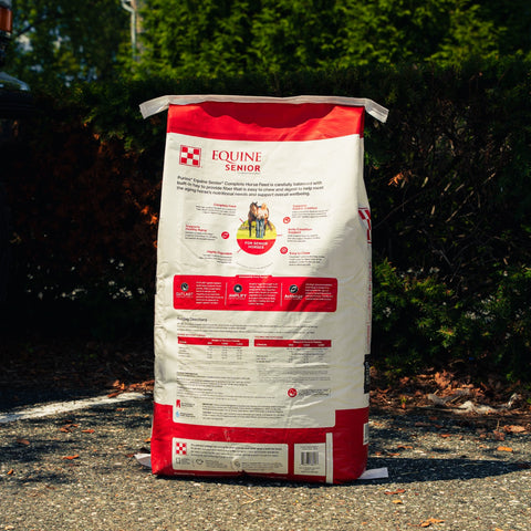 A large bag of Purina Equine Senior horse feed supplement stands outdoors on a gravel surface with dark green hedges in the background. The bag is white and red with a photo of a woman and horse on the front, and the Purina brand logo is clearly visible.