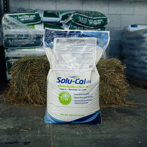 A large white bag of Solu-Cal USA Enhanced Calcium Products stands on a dark floor in front of stacked hay bales and other branded bags in a warehouse-like setting.