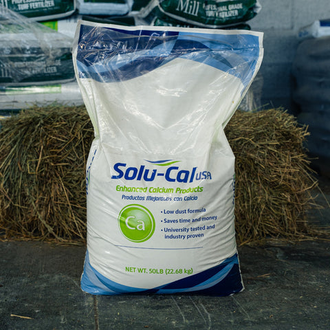 A 50 lb bag of Solu-Cal USA Enhanced Calcium Products standing upright on a concrete floor, with hay bales and other bags in the background.