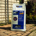 A large white bag of Lucerne Farms Hi Fiber Gold horse feed stands on a stone patio in front of a house with a wire fence and garden plants.