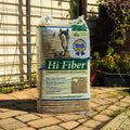 A large bag of Lucerne Farms Hi Fiber horse feed stands on a stone patio in front of a house with a wire fence and garden plants.