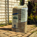 A large bag of Lucerne Farms Hi Fiber horse feed stands on a stone patio in front of a house with a wire fence and garden plants.