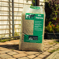 A large green bag of Lucerne Farms Alfa Supreme horse feed stands on a stone patio in front of a house with a wire fence and garden plants.