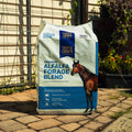 A large bag of Triple Crown Premium Alfalfa Forage Blend horse feed stands on a stone patio in front of a house with a wire fence and garden plants.