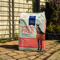 A large bag of Triple Crown Stress Free Forage Blend horse feed stands on a stone patio in front of a house with a wire fence and garden plants.