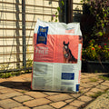A large bag of Triple Crown Stress Free Forage Blend horse feed stands on a stone patio in front of a house with a wire fence and garden plants.